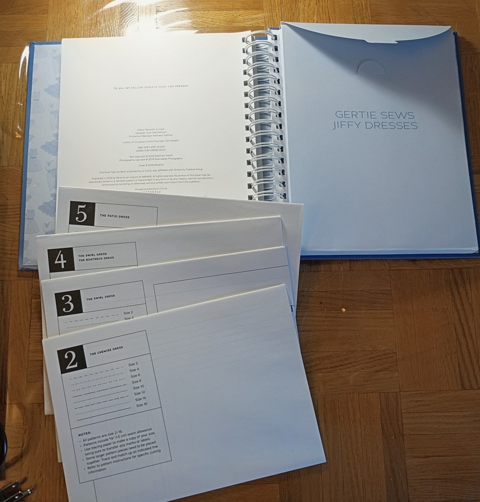A spiral-bound sewing book titled Gertie Sews Jiffy Dresses sitting open on a wooden table, with several separate white pattern sheets labeled with numbers 2 through 5 stacked neatly in the foreground.